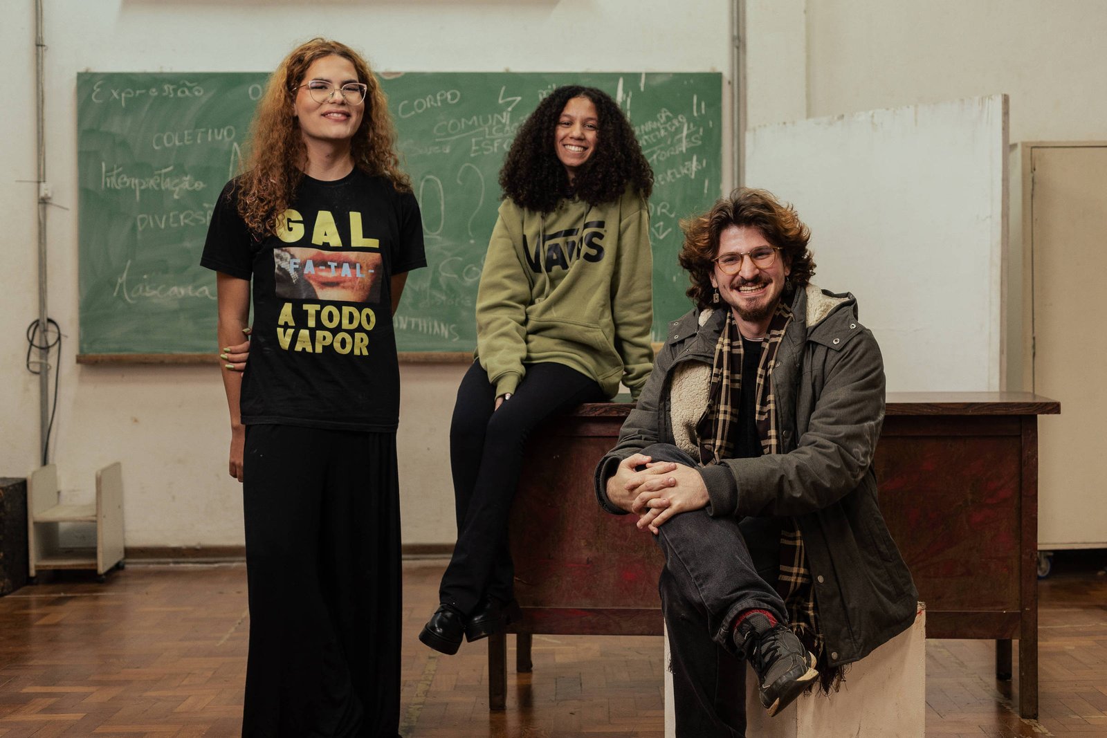 A imagem mostra três pessoas em uma sala de aula. À esquerda, uma mulher com cabelo cacheado e óculos usa uma camiseta preta com a frase