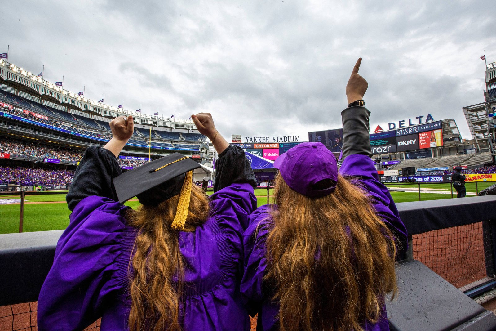 Duas pessoas de costas, vestindo togas de formatura roxas e chapéus, estão levantando os braços em celebração em direção ao campo do Yankee Stadium. O céu está nublado e o estádio está cheio de pessoas. Ao fundo, há uma tela com o nome