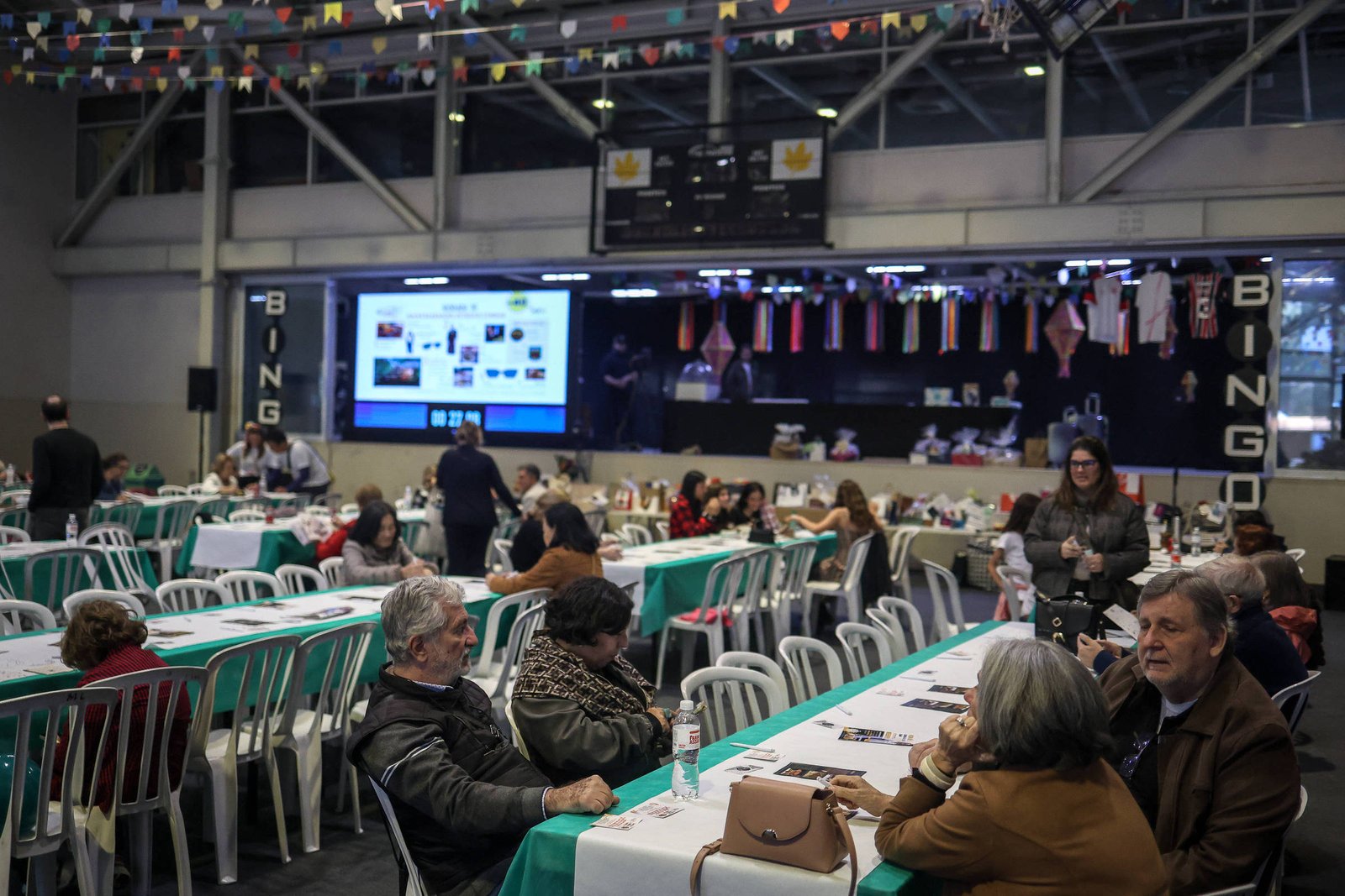 A imagem mostra um grande salão de eventos com mesas dispostas em fileiras. Algumas pessoas estão sentadas nas mesas, conversando e interagindo. Ao fundo, há um palco com uma tela exibindo imagens e bandeiras de diferentes países penduradas. O ambiente é decorado com bandeirinhas coloridas, criando uma atmosfera festiva. A iluminação é suave e o espaço parece estar bem frequentado.