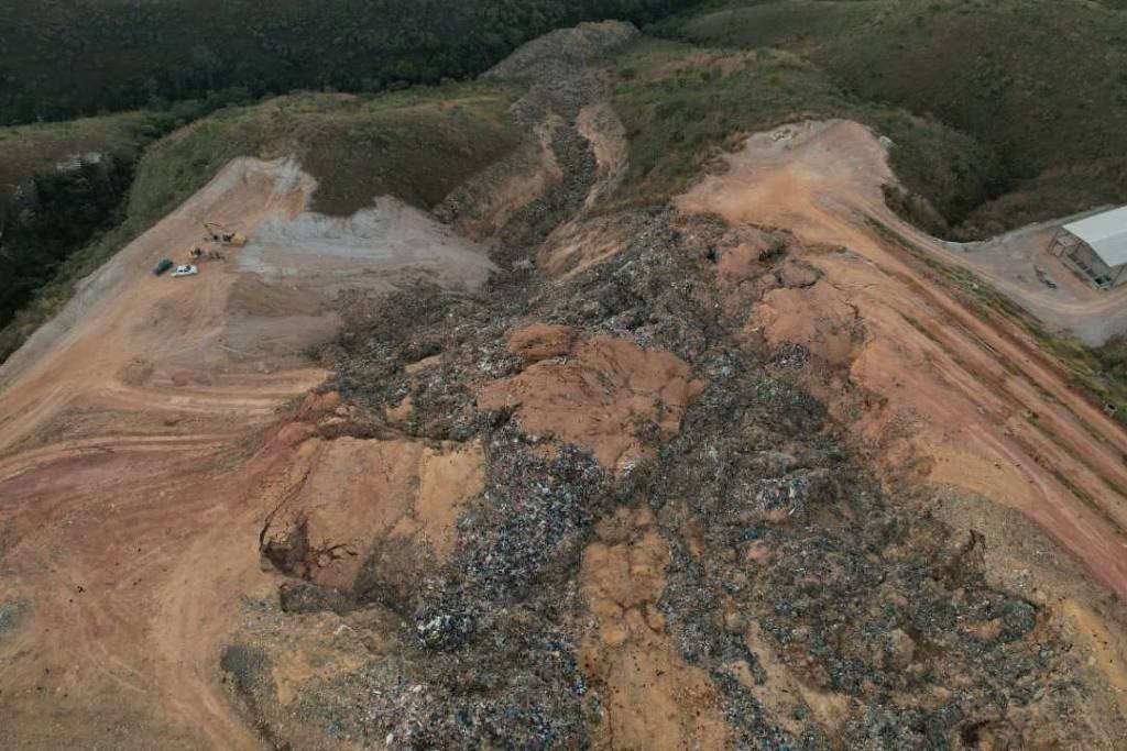 A imagem mostra um aterro sanitário com uma grande quantidade de lixo acumulado em uma área montanhosa. O terreno é irregular, com uma mistura de solo exposto e resíduos visíveis. Ao fundo, há uma vegetação densa e uma estrutura de metal, possivelmente um galpão, localizada em uma área mais limpa do aterro. A cena é capturada de uma perspectiva aérea, evidenciando a extensão do aterro e a disposição dos resíduos.