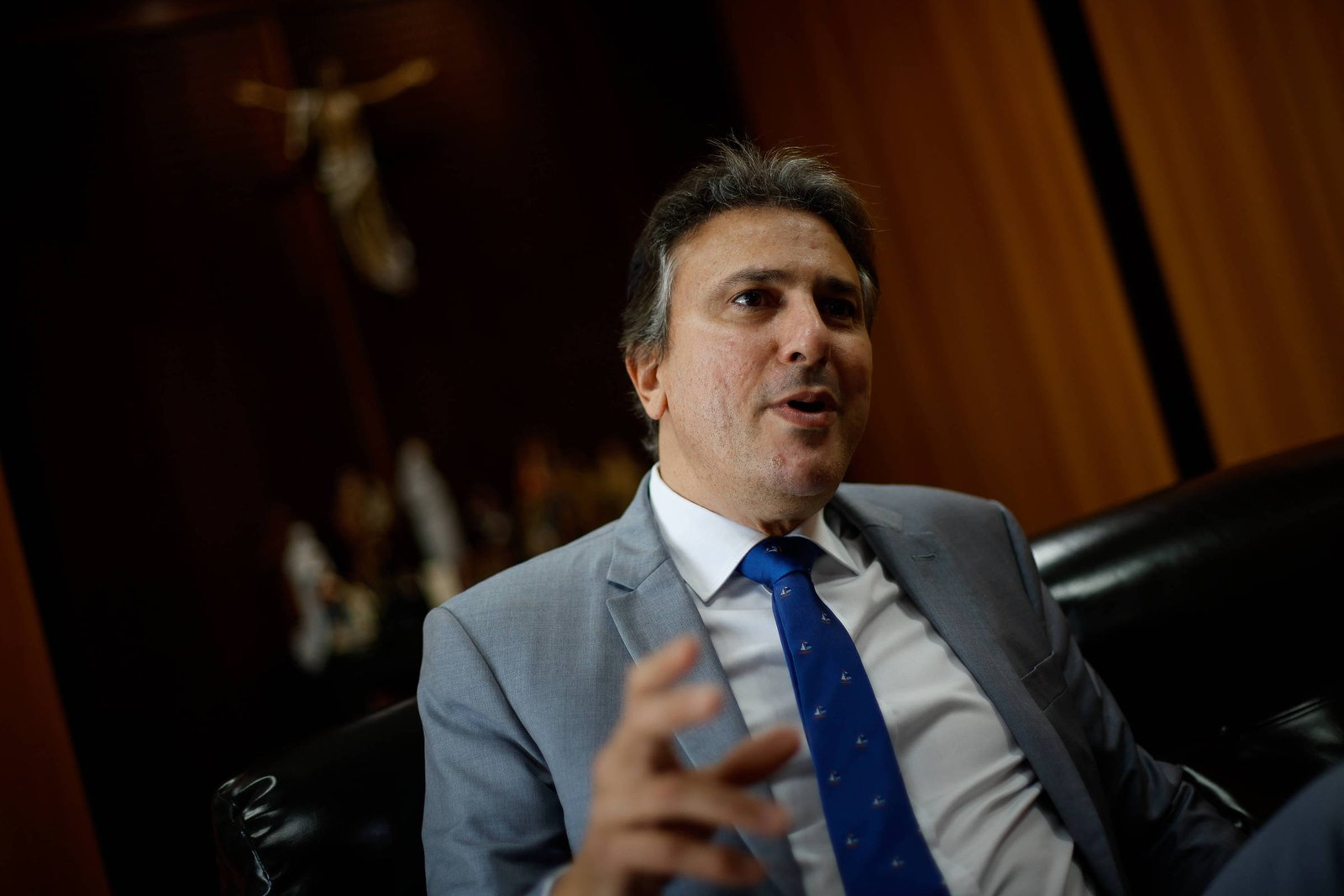 Um homem de terno cinza e gravata azul com bolinhas brancas está sentado em um sofá preto. Ele está gesticulando com a mão direita enquanto fala. Ao fundo, há uma parede de madeira e uma figura religiosa visível, possivelmente uma cruz. O ambiente parece ser um escritório ou sala de reuniões.