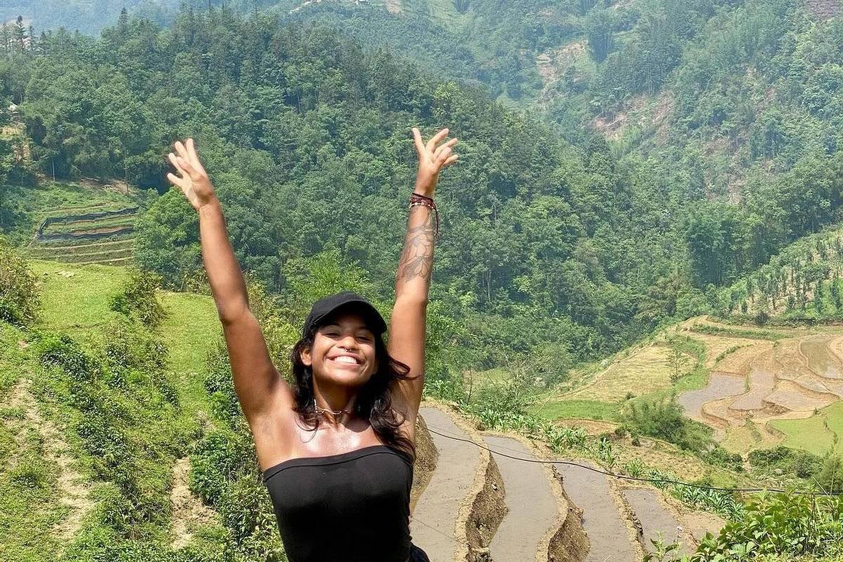 Uma mulher está em uma área montanhosa, com as mãos levantadas em sinal de alegria. Ela usa uma blusa preta e shorts pretos. Ao fundo, há uma paisagem verde com montanhas e campos de arroz em terraços. O céu está claro, com algumas nuvens.