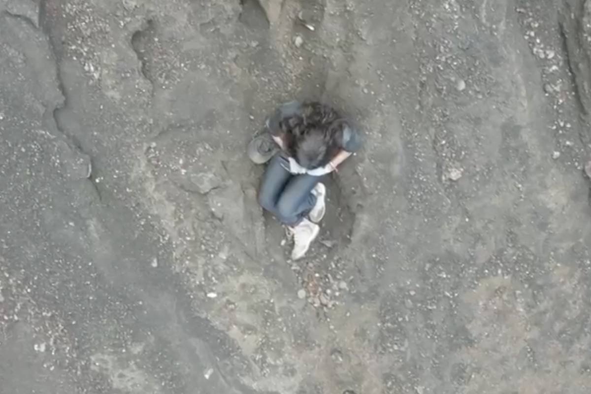 A imagem mostra uma pessoa sentada em uma área rochosa e irregular, com o corpo encolhido e as pernas dobradas. A pessoa está vestindo uma camiseta e jeans, e parece estar em uma posição de reflexão ou descanso. O solo ao redor é composto por pedras e terra, com algumas pequenas pedras soltas visíveis.