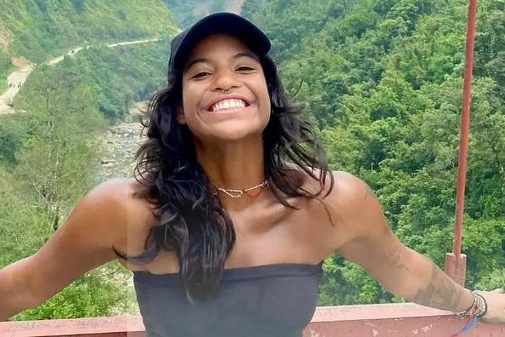 Uma mulher sorridente está em uma ponte, com os braços abertos, em um cenário natural exuberante. Ao fundo, há montanhas verdes e um rio visível. A mulher usa uma blusa preta e um boné. O ambiente parece ser ensolarado e alegre, transmitindo uma sensação de liberdade e felicidade.