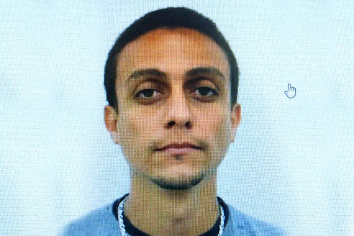 A imagem mostra um homem com cabelo curto e escuro, olhando diretamente para a câmera. Ele tem uma expressão neutra e está vestindo uma camiseta escura. O fundo da imagem é branco, e há uma leve sombra ao redor do rosto. O homem tem uma corrente visível em seu pescoço e um leve traço de barba.