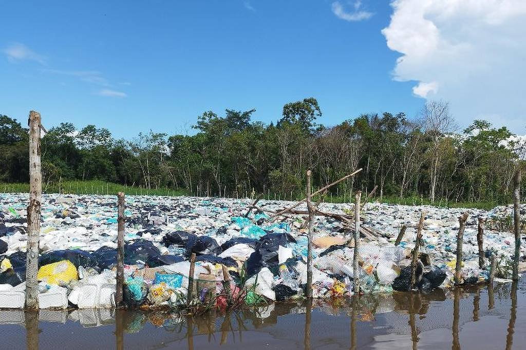 A imagem mostra uma grande quantidade de lixo acumulado em uma área alagada, com sacos plásticos e outros resíduos flutuando na água. Ao fundo, há uma vegetação densa, composta por árvores e arbustos, sob um céu azul com algumas nuvens brancas. A cena retrata um problema ambiental significativo.