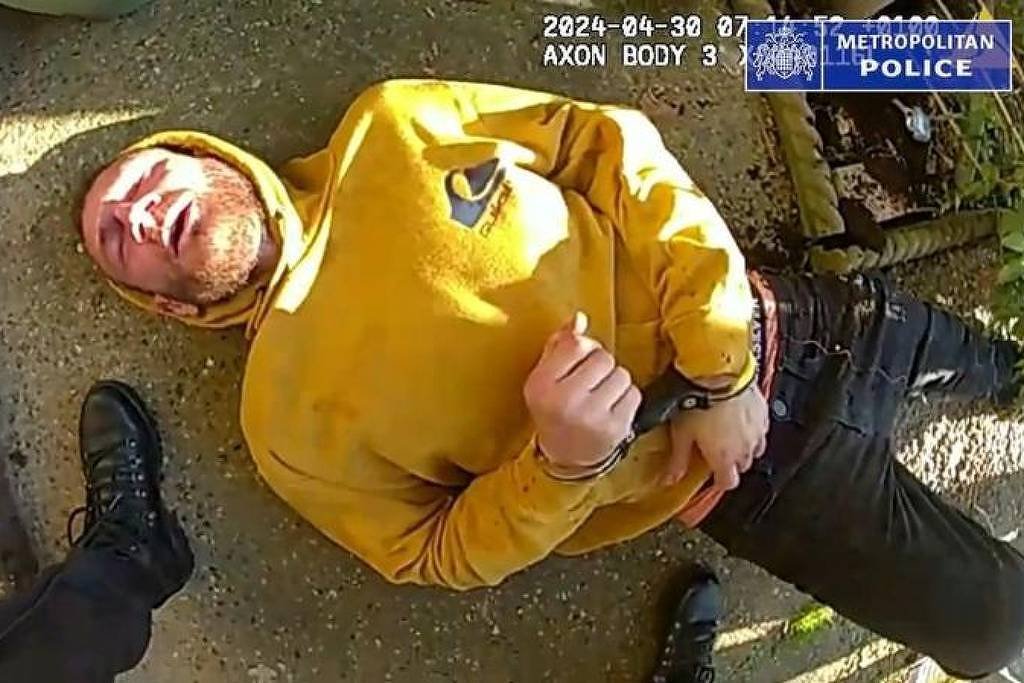 A imagem mostra um homem deitado no chão, vestindo um moletom amarelo. Ele está algemado e parece estar em uma posição de rendição, com uma expressão facial que sugere desconforto ou dor. Um pé, possivelmente de um policial, está visível próximo ao homem. O fundo é um ambiente externo, com luz natural.