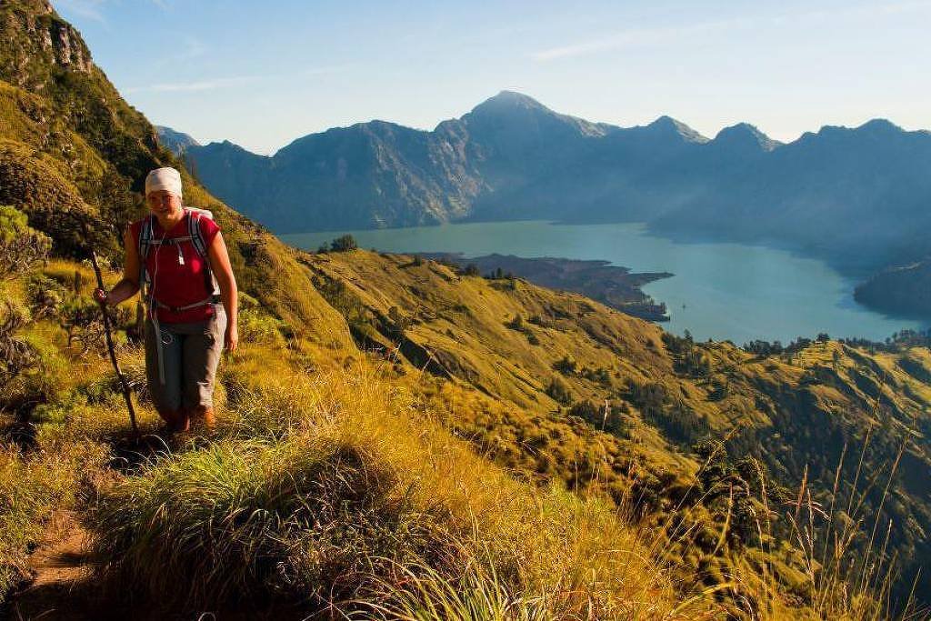 Uma pessoa está caminhando por um trilha em uma montanha, cercada por vegetação alta e exuberante. Ao fundo, há um lago e montanhas adicionais, com um céu claro e ensolarado. A pessoa usa uma camiseta vermelha e shorts, segurando um bastão de caminhada.