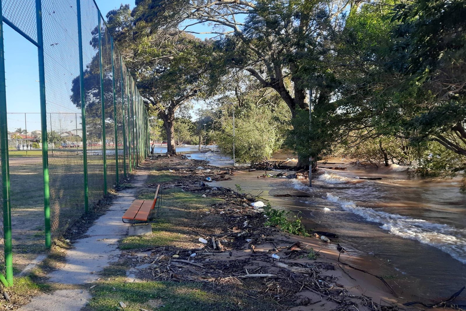 A imagem mostra uma área de recreação ao ar livre, com uma quadra de esportes ao fundo cercada por uma grade verde. À esquerda, há um banco de madeira e, à direita, um rio que transbordou, inundando a margem. A vegetação ao redor é densa, com árvores grandes e folhagens. O céu está claro e ensolarado, indicando um dia bonito, apesar da inundação visível.