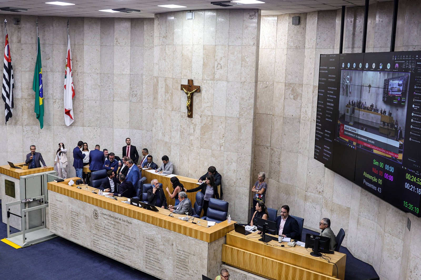 A imagem mostra o interior de uma câmara legislativa. Há várias pessoas sentadas em cadeiras azuis, algumas em pé, e um homem falando ao microfone. Ao fundo, há uma parede de pedra clara com uma cruz, bandeiras do Brasil e do estado de São Paulo, e um grande painel eletrônico exibindo informações sobre a sessão. O ambiente é bem iluminado e formal.