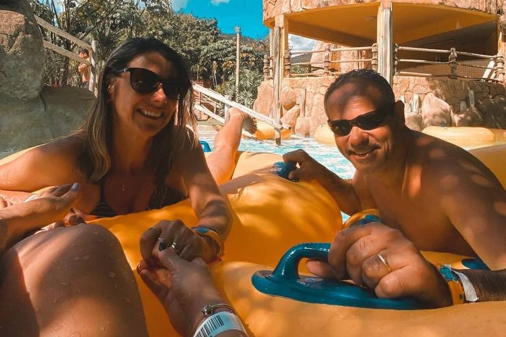 A imagem mostra um grupo de quatro pessoas em um escorregador de água em um parque aquático. Elas estão em um bote amarelo, sorrindo e se divertindo. Ao fundo, há uma estrutura de escorregador e árvores. O céu está claro e azul. Na parte superior da imagem, há um texto que diz:
