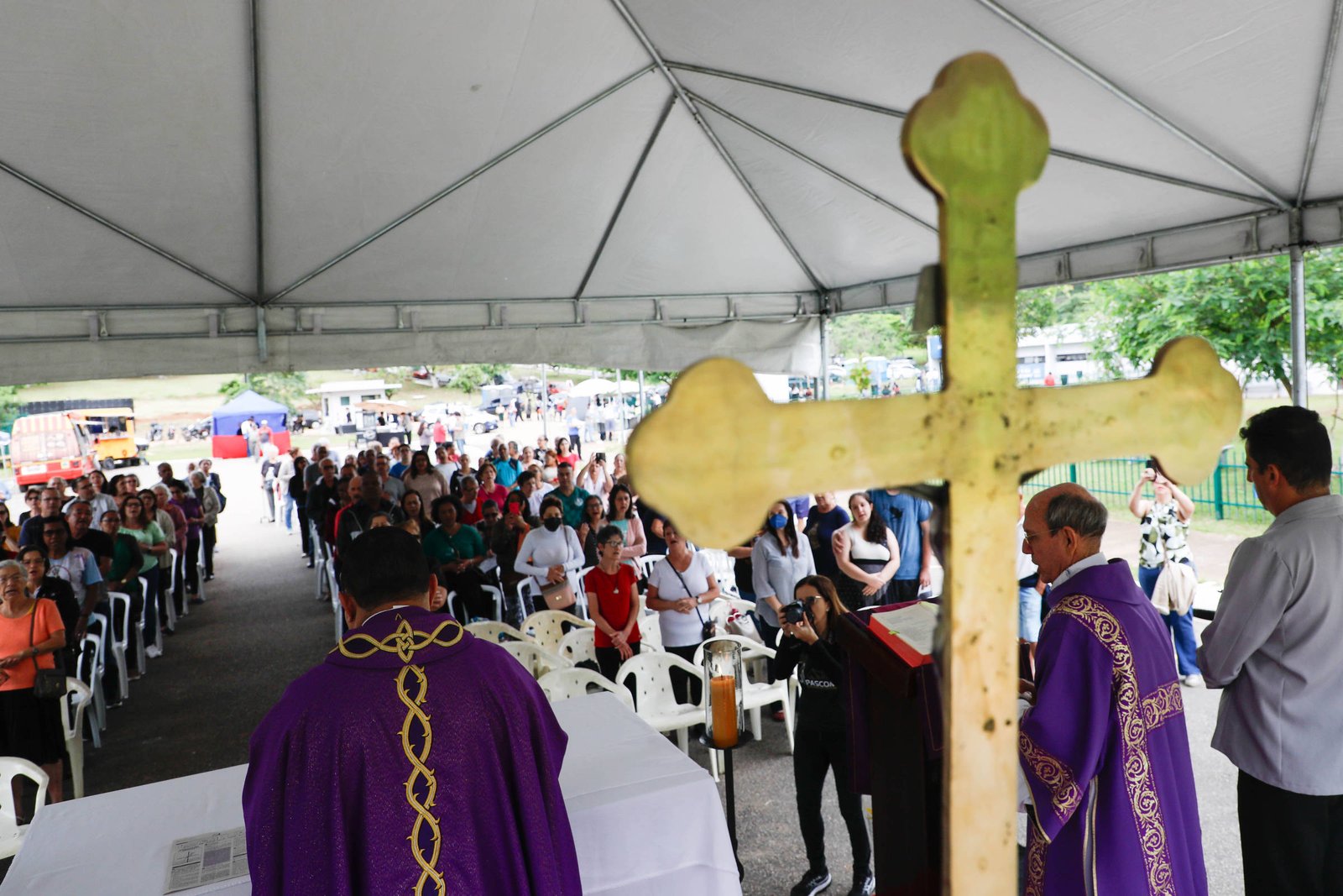 A imagem mostra uma celebração religiosa ao ar livre, sob uma tenda. Em primeiro plano, há uma cruz grande e dourada. Ao fundo, um grupo de pessoas está assistindo à cerimônia, com algumas sentadas em cadeiras brancas. Dois sacerdotes estão visíveis, um deles de costas, usando vestes litúrgicas roxas. O ambiente é festivo e há uma atmosfera de comunhão entre os participantes.