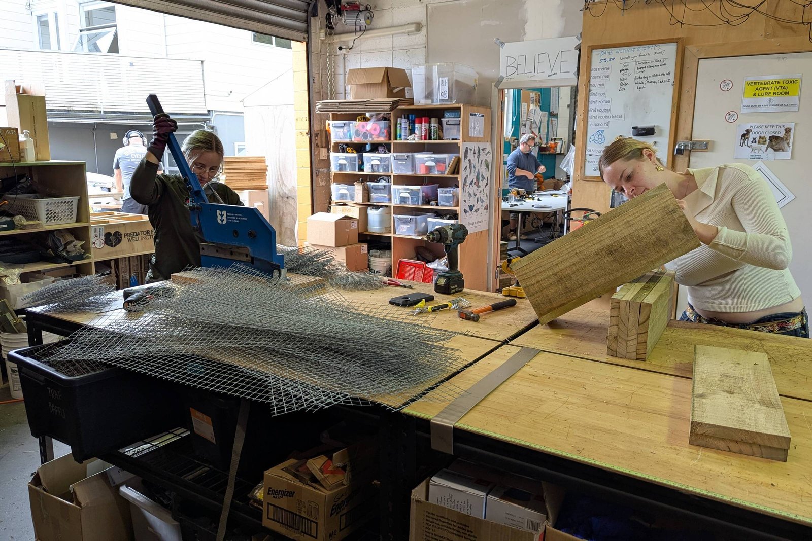 A imagem mostra uma oficina com duas pessoas trabalhando em uma mesa. À esquerda, uma mulher está usando uma ferramenta para cortar uma grade de metal, enquanto à direita, outra mulher está segurando uma peça de madeira. Ao fundo, há prateleiras com materiais e ferramentas organizadas. A iluminação é clara e o ambiente parece ser um espaço de trabalho colaborativo.