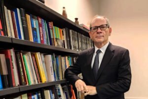 Um homem de terno preto e gravata, com óculos, está posando ao lado de uma estante cheia de livros. Ele está com os braços cruzados e um leve sorriso no rosto. O ambiente é bem iluminado, com uma parede clara e um piso azul. Ao fundo, há uma mesa de escritório e mais livros na estante.