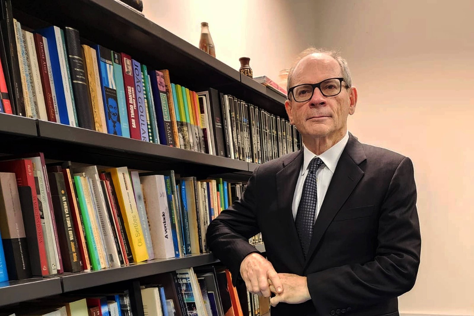 Um homem de terno preto e gravata, com óculos, está posando ao lado de uma estante cheia de livros. Ele está com os braços cruzados e um leve sorriso no rosto. O ambiente é bem iluminado, com uma parede clara e um piso azul. Ao fundo, há uma mesa de escritório e mais livros na estante.