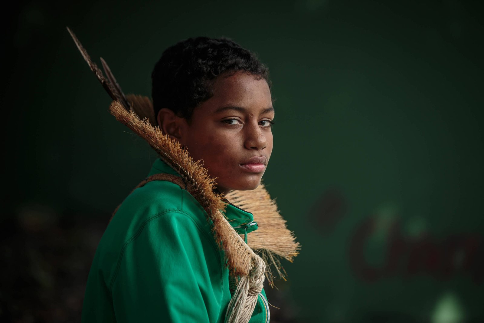 Um jovem com cabelo curto e encaracolado está posando para a foto. Ele usa uma camisa verde e tem uma expressão séria. Nas costas, ele carrega um cocar feita de palha. O fundo é de cor verde escura, criando um contraste com a roupa do jovem.