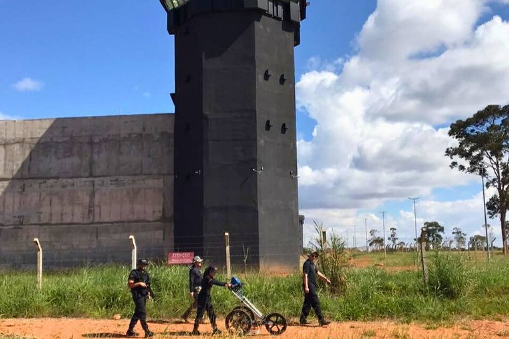 A imagem mostra uma torre de vigilância de um presídio, que é alta e de cor preta, com janelas na parte superior. Ao lado da torre, há um muro de concreto. Na frente, três pessoas vestidas com roupas escuras estão caminhando, uma delas empurrando um carrinho. O céu está claro com algumas nuvens e há vegetação ao redor.