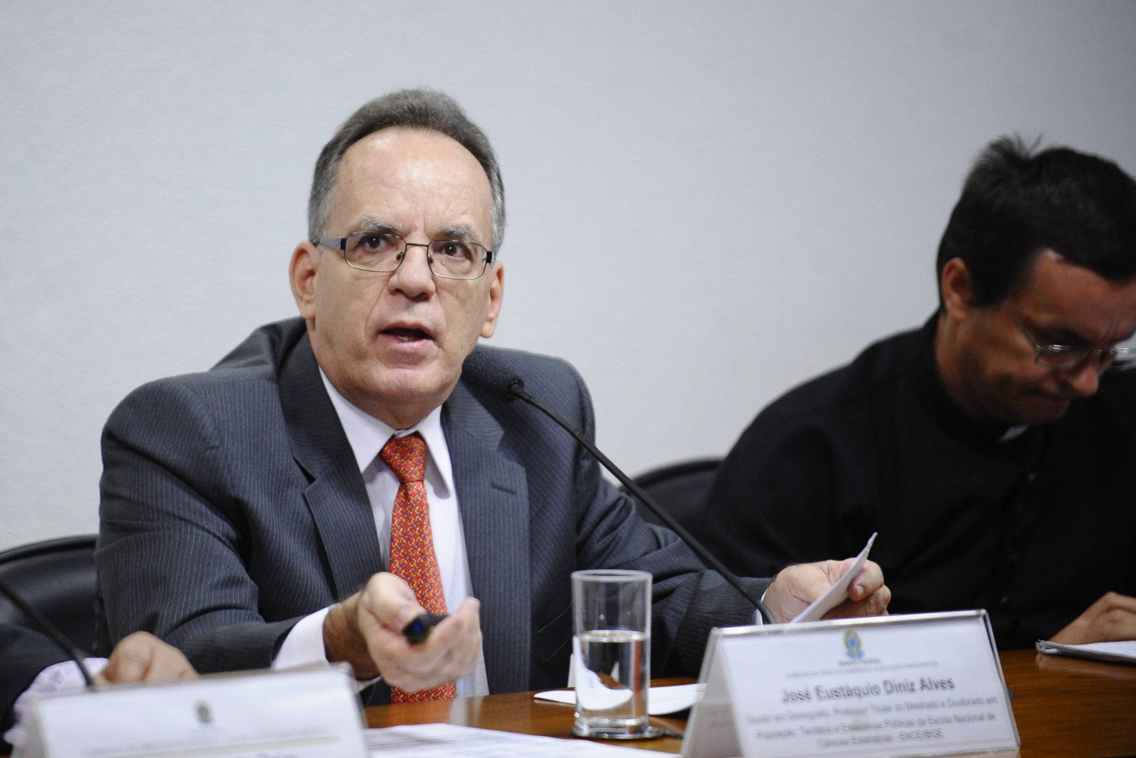 Um homem de óculos e terno cinza, com uma gravata vermelha, está falando em uma mesa de reunião. Ele parece estar explicando algo, gesticulando com a mão. À sua frente, há um copo de água e papéis. Ao fundo, um outro homem, vestido de preto, está sentado e prestando atenção. O ambiente é de uma sala de reuniões, com paredes claras e uma mesa de madeira.