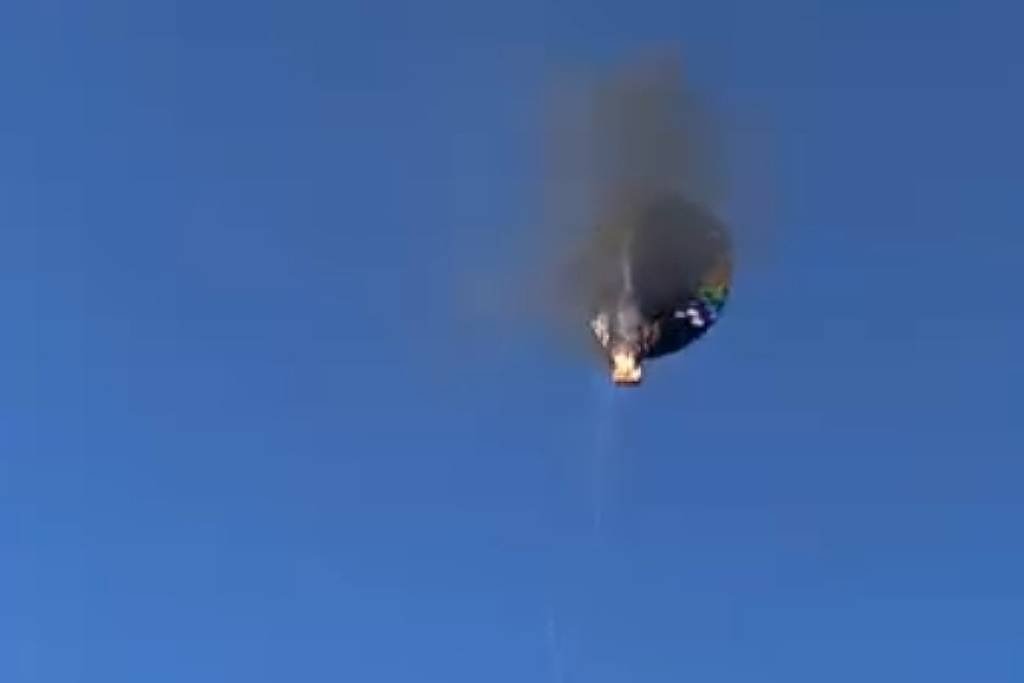 A imagem mostra um balão de ar quente no céu, com fumaça saindo de sua parte superior. O céu é azul e claro, e o balão parece estar em uma posição elevada, com uma parte colorida visível. A fumaça indica que o balão pode estar pegando fogo ou passando por algum tipo de falha.