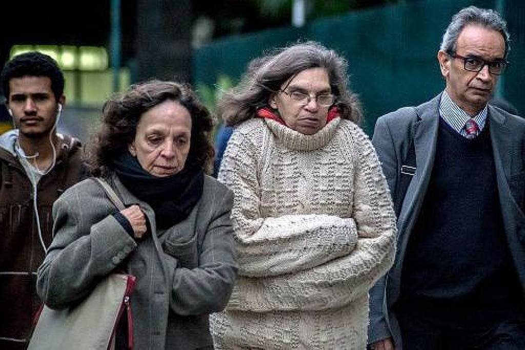 A imagem mostra um grupo de quatro pessoas caminhando em uma área urbana. Duas mulheres estão à frente, uma delas vestindo um casaco escuro e a outra um suéter claro, ambas com expressões sérias. Um homem de óculos e terno caminha ao lado delas, enquanto um jovem atrás usa um casaco marrom. O ambiente parece ser de uma cidade, com uma grade verde ao fundo.