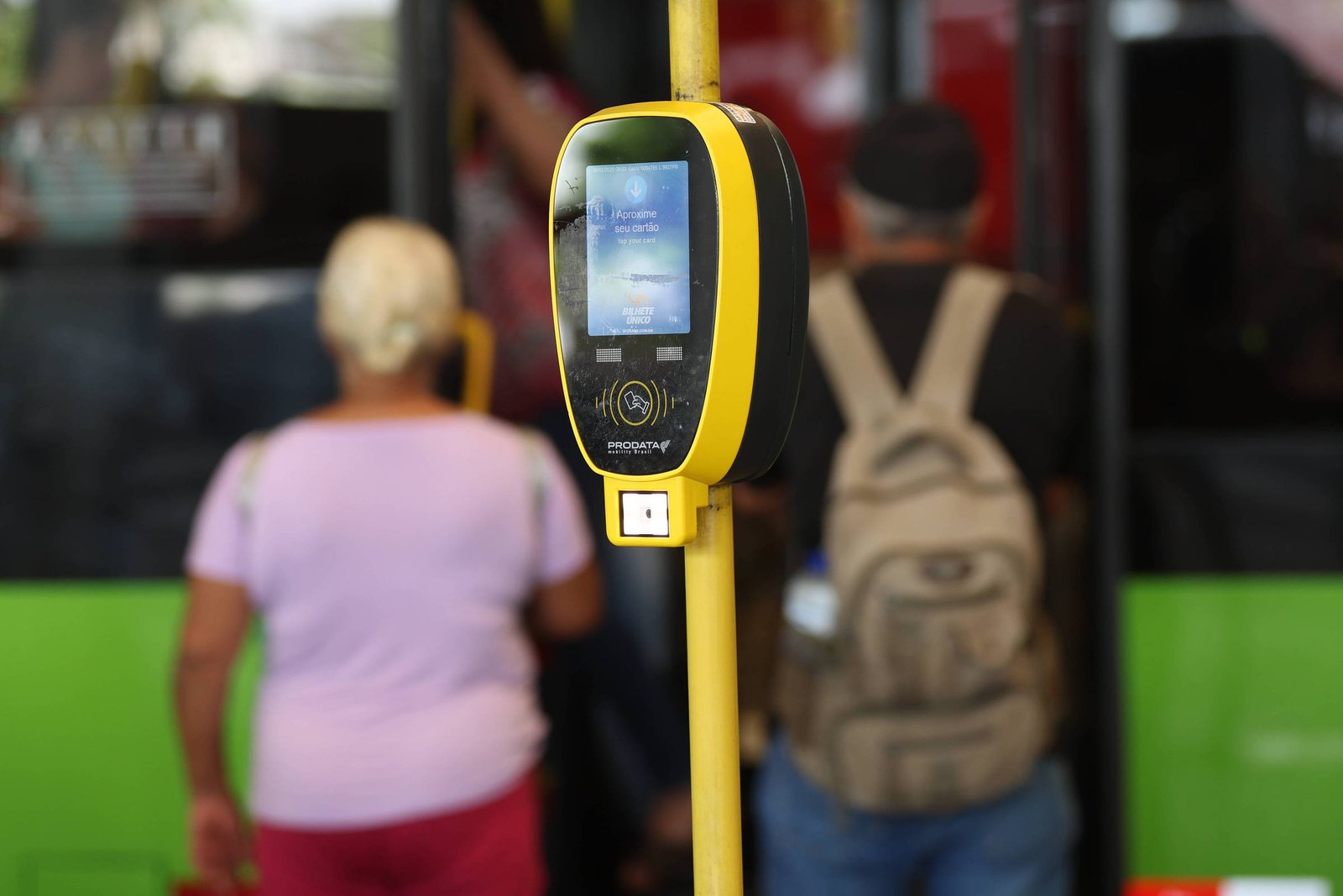 A imagem mostra um validador de bilhete único em uma parada de ônibus. O dispositivo é amarelo e preto, com uma tela visível. Ao fundo, algumas pessoas estão entrando no ônibus, uma delas é uma mulher de cabelo curto e grisalho, vestindo uma blusa rosa. Outra pessoa, com uma mochila, está de costas, enquanto um homem com um boné também é visível. O ônibus é de cor verde.