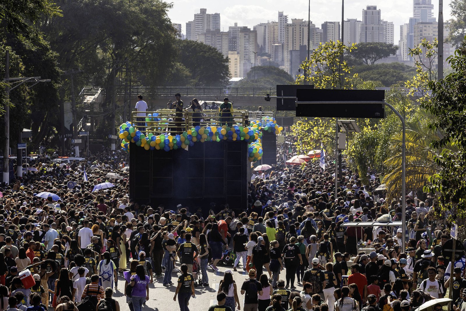 A imagem mostra uma grande multidão reunida em um evento ao ar livre, com pessoas de várias idades e estilos. No centro, há um caminhão de som decorado com balões coloridos, onde estão duas pessoas em cima, possivelmente se apresentando. Ao fundo, é possível ver uma cidade com prédios altos e árvores ao redor. O céu está claro e ensolarado, e algumas pessoas estão usando guarda-sóis.