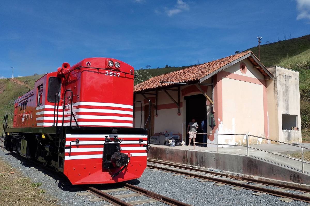 A imagem mostra um trem vermelho com listras brancas estacionado em uma plataforma de uma estação. Ao fundo, há um edifício de estação com paredes em tom claro e um telhado de telhas. Algumas pessoas estão próximas à entrada da estação. O céu está claro e azul, com algumas nuvens.