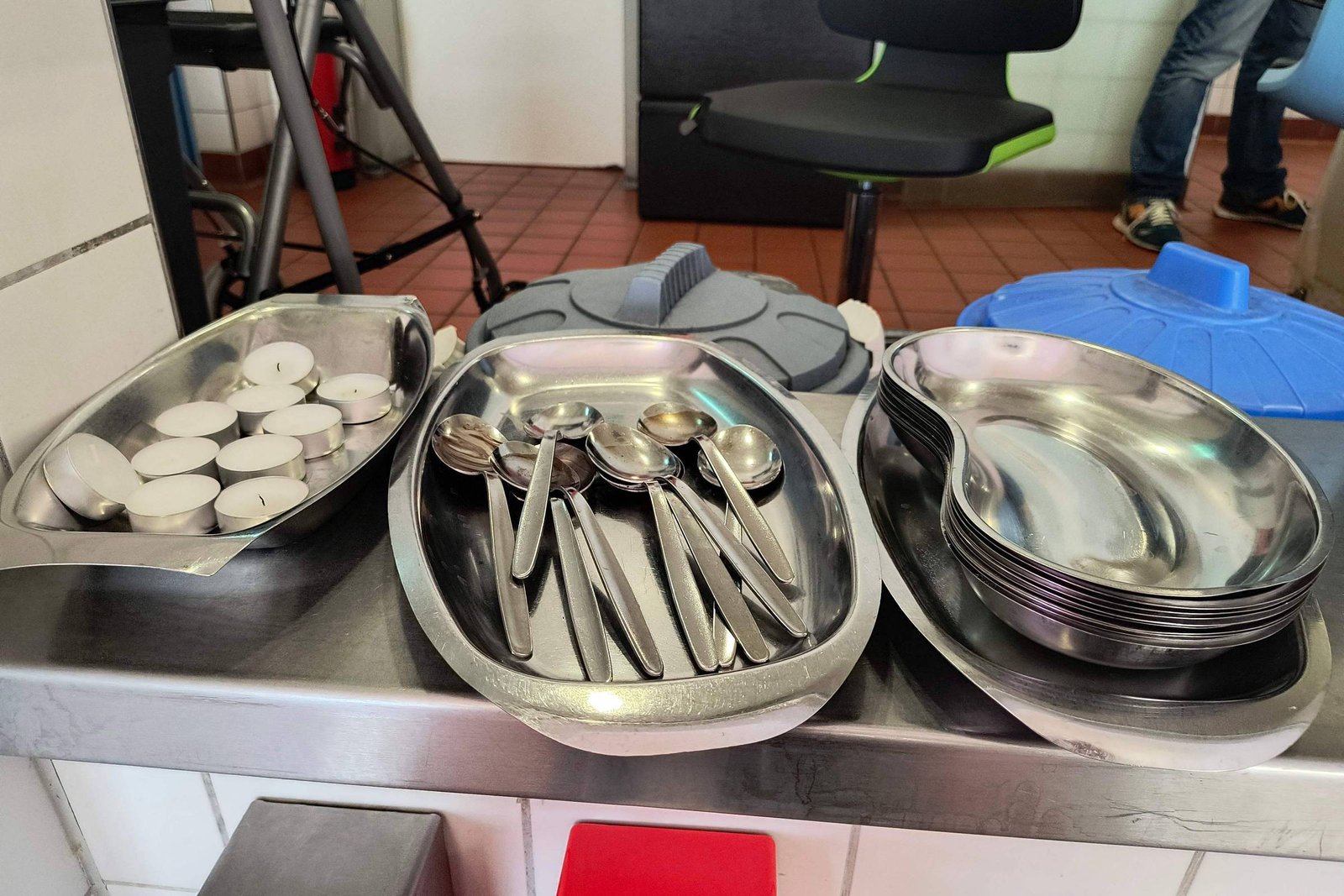A imagem mostra uma mesa com utensílios de cozinha. Há bandejas de metal com colheres e pratos empilhados. Também estão presentes algumas velas em copos pequenos. Ao fundo, é possível ver uma cadeira e parte de uma mesa.