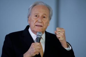 Um homem de cabelos grisalhos e pele clara está falando ao microfone. Ele usa um terno escuro e uma gravata laranja. O homem está levantando a mão em um gesto de ênfase, com o punho cerrado. O fundo é neutro e desfocado, sugerindo um ambiente de apresentação ou discurso.