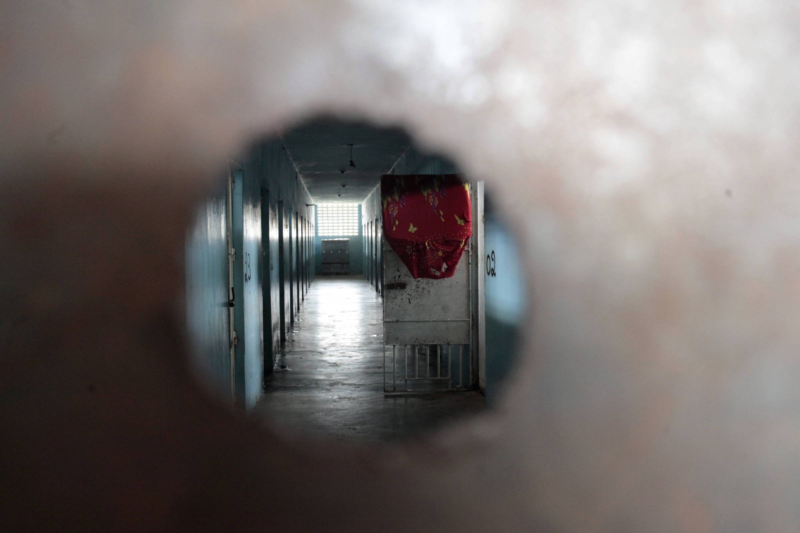 A imagem mostra uma visão através de um buraco em uma superfície metálica. No fundo, é possível ver um corredor iluminado, com paredes claras e um piso reflexivo. Há um objeto vermelho pendurado em uma das paredes, e a luz entra pela extremidade do corredor, criando um contraste com a escuridão ao redor do buraco.