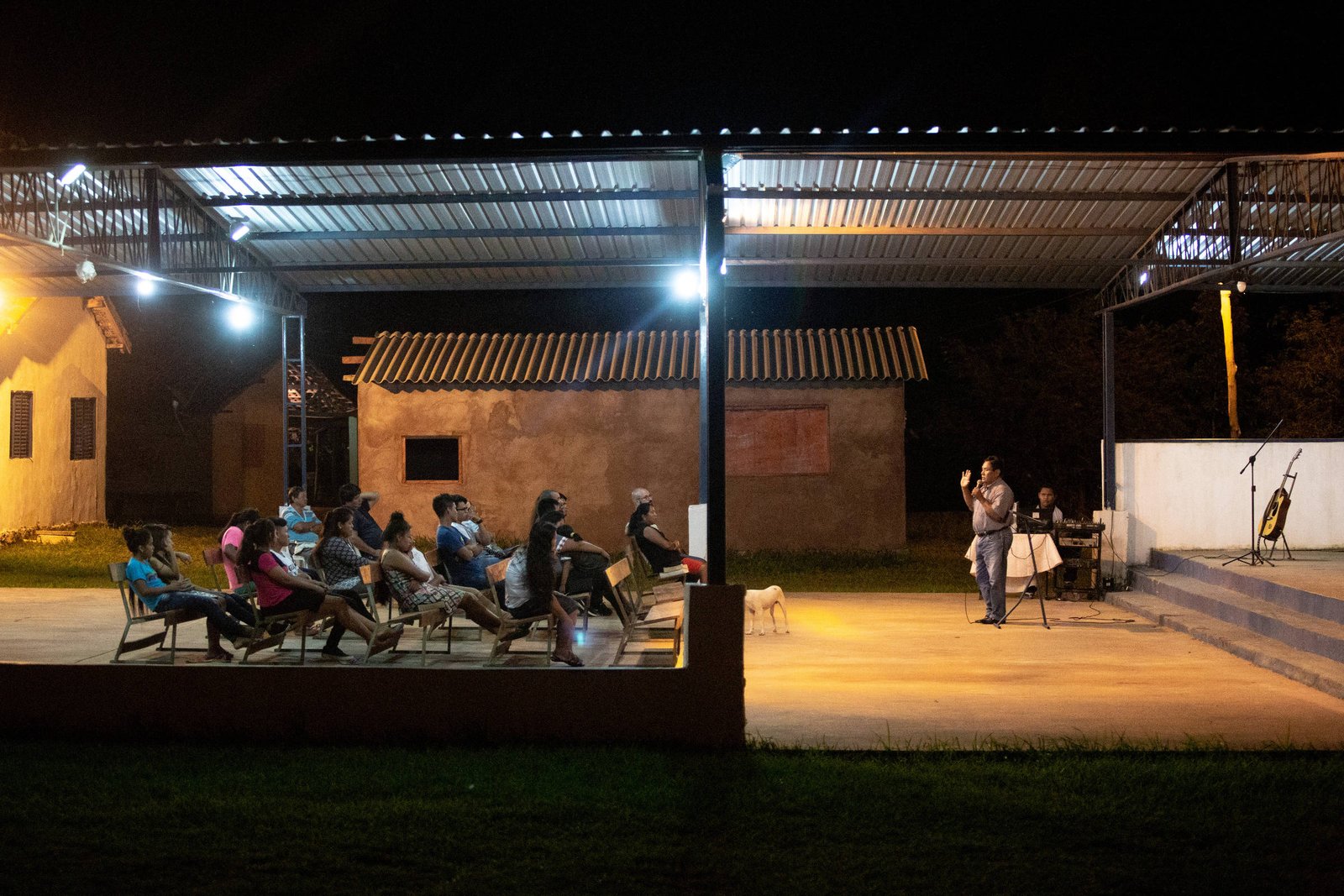 A imagem mostra um evento noturno em um espaço aberto. Há um grupo de pessoas sentadas em cadeiras, assistindo a uma apresentação que ocorre em um palco à direita. O ambiente é iluminado por luzes artificiais, e ao fundo, há uma construção simples. O céu está escuro, indicando que é noite.