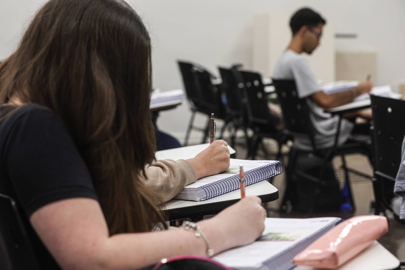 A imagem mostra dois estudantes em uma sala de aula. À esquerda, uma estudante com cabelo longo e escuro está escrevendo em um caderno, enquanto à direita, um estudante com óculos e cabelo curto está sentado em uma cadeira, também escrevendo. As mesas são brancas e há cadernos e materiais escolares visíveis sobre elas. O ambiente parece ser bem iluminado e organizado.