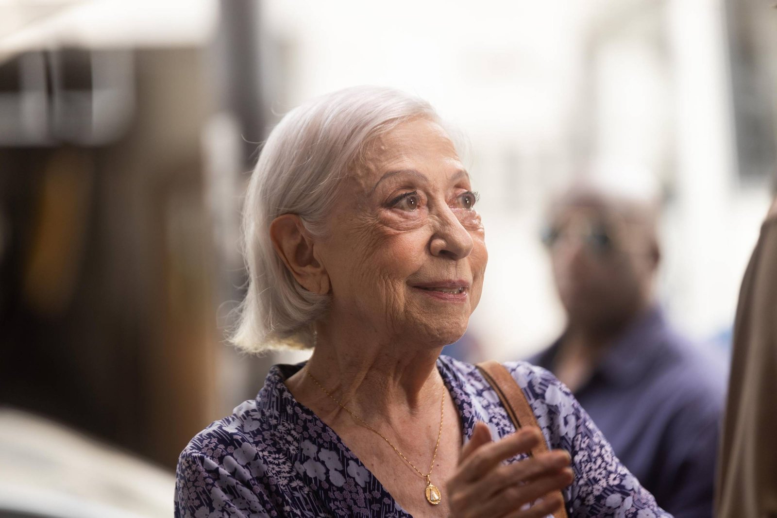 A imagem mostra uma mulher idosa com cabelo curto e grisalho, usando uma blusa com estampas em tons de azul. Ela está sorrindo e parece estar em uma conversa, com as mãos levantadas em um gesto de entusiasmo. Ao fundo, há uma pessoa com óculos escuros e uma camisa escura, mas seu rosto não é claramente visível.