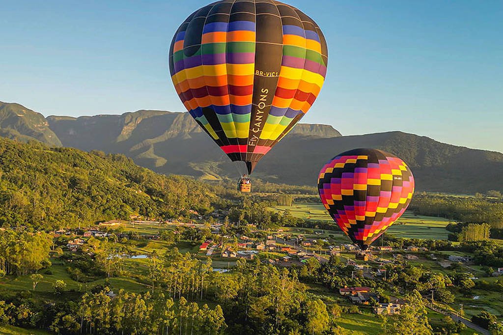 A imagem mostra dois balões de ar quente coloridos flutuando sobre uma paisagem verdejante. O balão da esquerda é predominantemente preto com listras coloridas, enquanto o balão da direita é mais vibrante, com um padrão de cores em rosa, amarelo e verde. Abaixo, há uma pequena vila com casas e árvores, e ao fundo, montanhas cobertas de vegetação sob um céu claro.