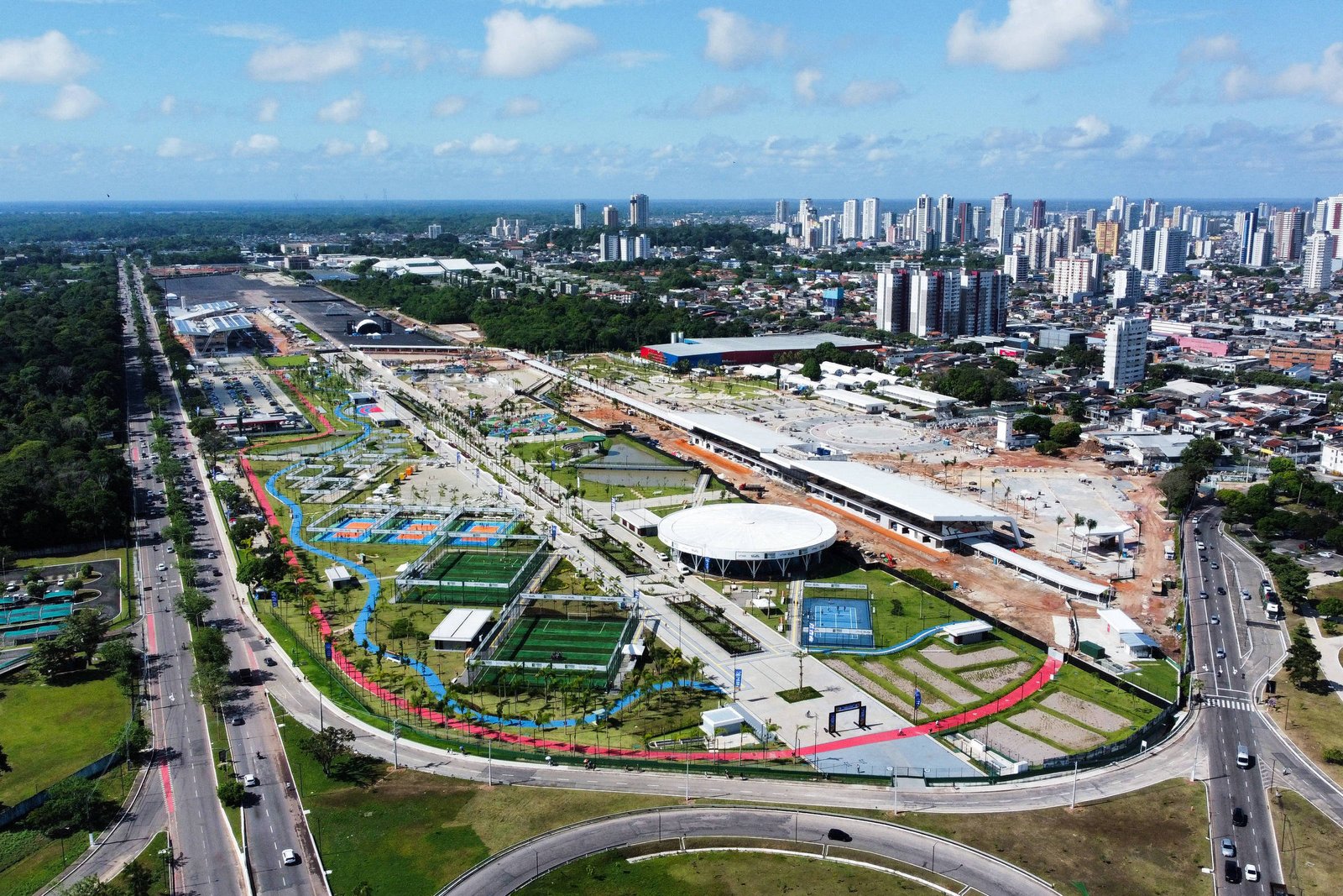 A imagem mostra uma vista aérea de uma área urbana em desenvolvimento, com várias construções e um grande espaço verde. Há estradas que cercam a área, com um padrão de caminhos coloridos e áreas de lazer visíveis. Ao fundo, pode-se ver uma cidade com prédios altos e um céu parcialmente nublado.