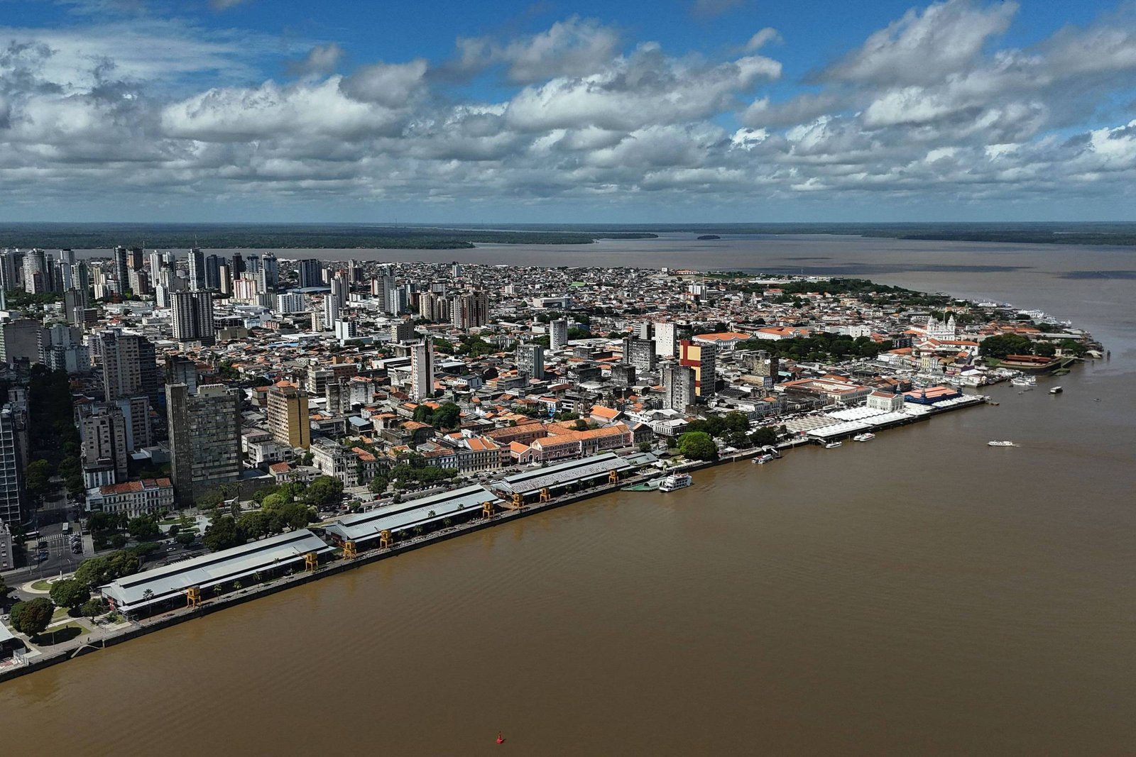 A imagem mostra uma vista aérea de uma cidade costeira, com prédios altos e uma área urbana densa à esquerda. À direita, há um rio de cor marrom, com algumas embarcações visíveis. O céu está parcialmente nublado, com nuvens brancas e um fundo azul. A cidade parece estar localizada em uma região tropical, com vegetação ao longo da costa.