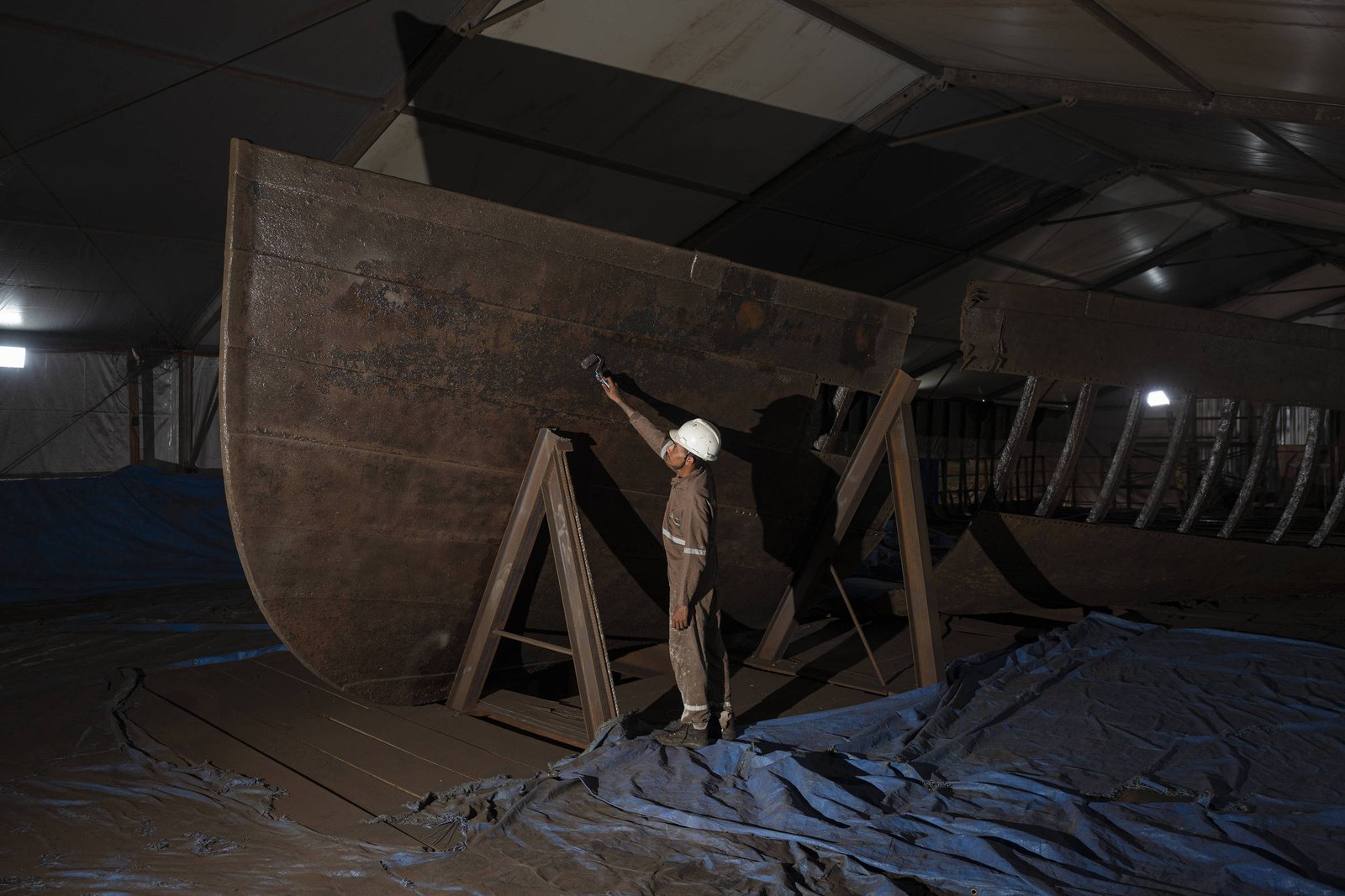 Um trabalhador está em um estaleiro naval, embaixo de uma tenda, realizando um trabalho de acabamento em uma grande estrutura de metal que parece ser a proa de um barco. Ele está usando um capacete de segurança e segurando uma ferramenta, enquanto a estrutura metálica está apoiada em suportes. O chão está coberto com uma lona azul e há iluminação artificial ao fundo.