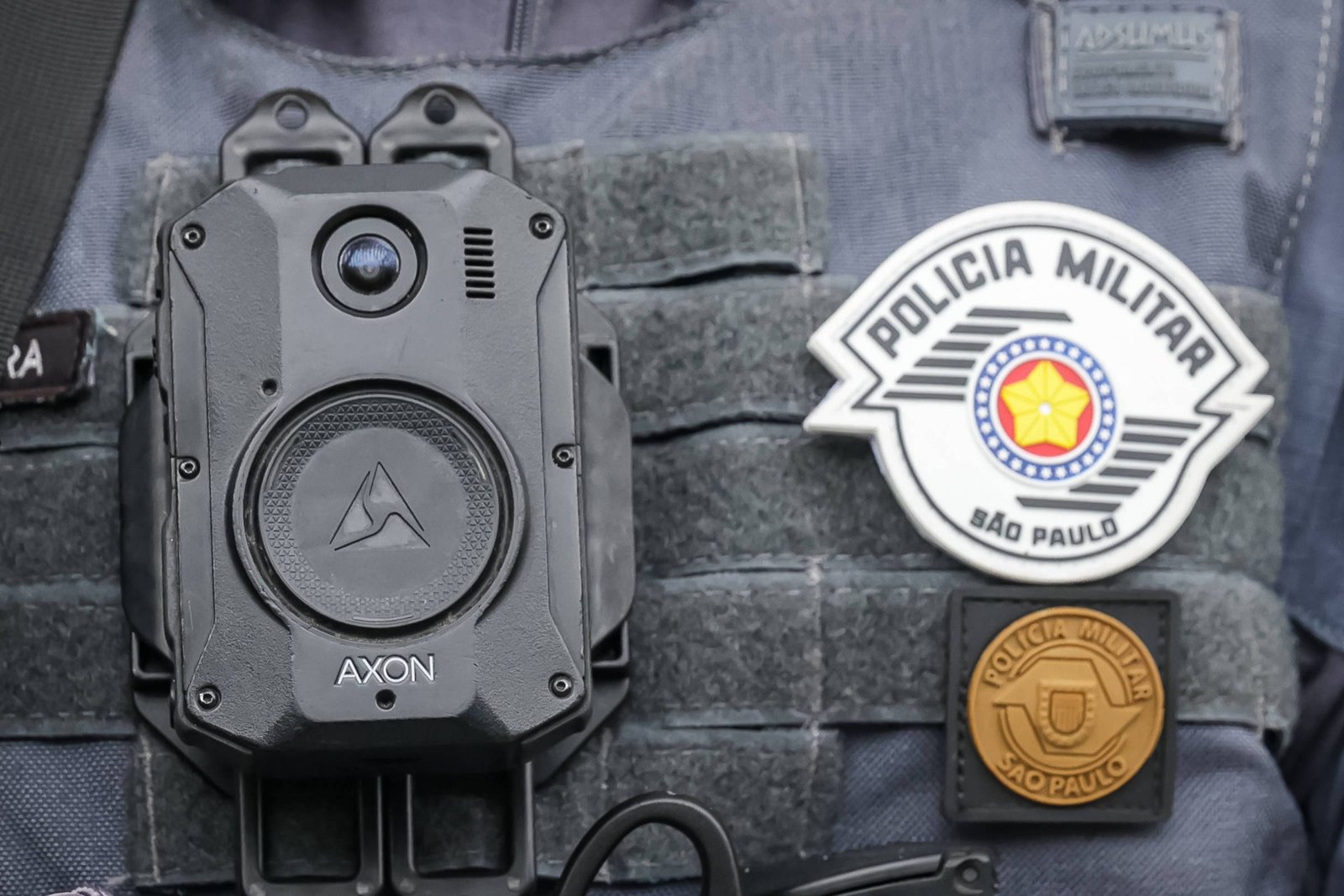 A imagem mostra um colete tático de um policial, com uma câmera de corpo (body cam) presa na parte frontal. A câmera é preta e possui o logotipo da marca