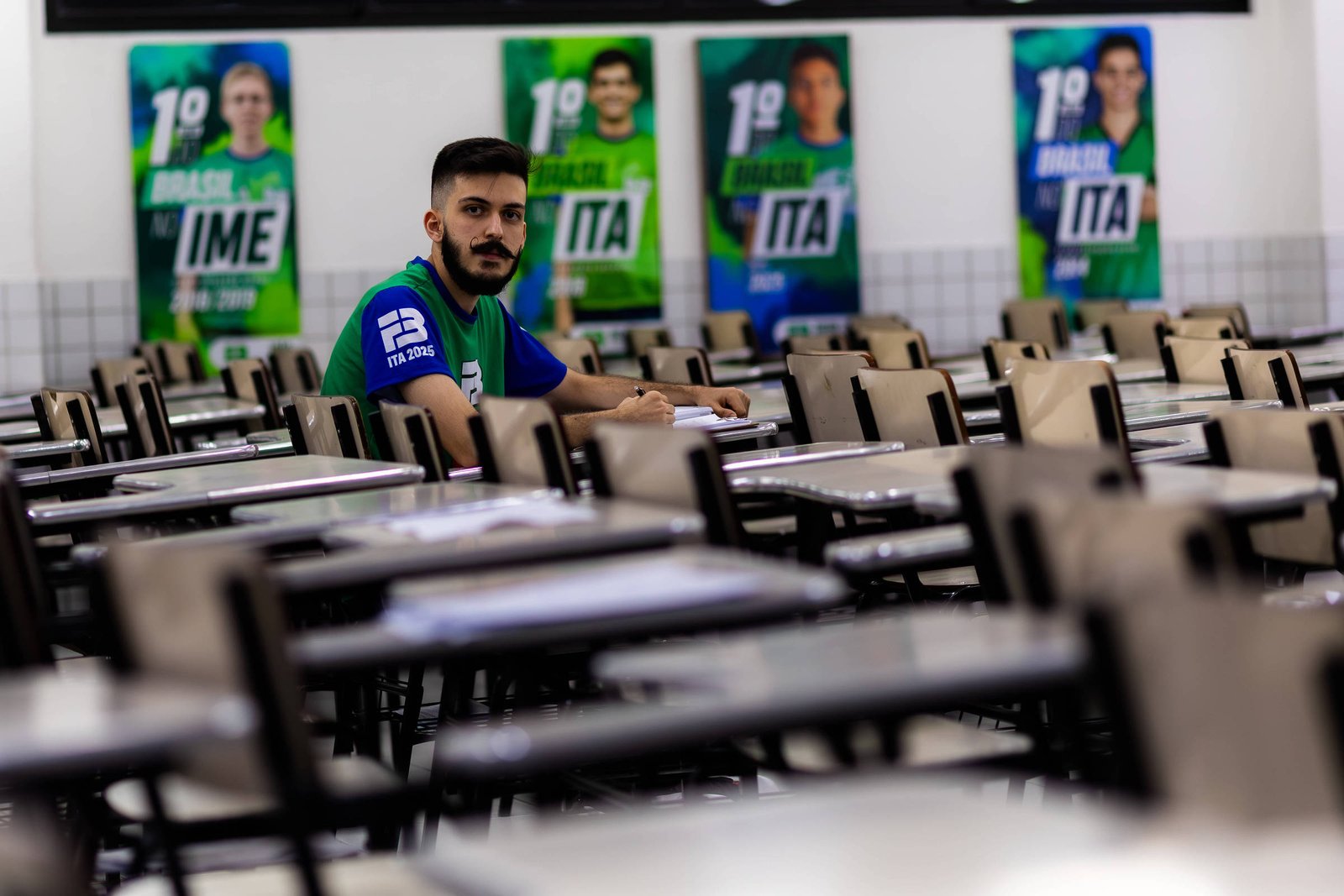 A imagem mostra um jovem sentado em uma mesa em um refeitório vazio. Ele está usando uma camiseta azul e parece estar escrevendo em um caderno. Ao fundo, há cartazes com os números 1 e 2, que parecem se referir a competições ou eventos, e estão relacionados a