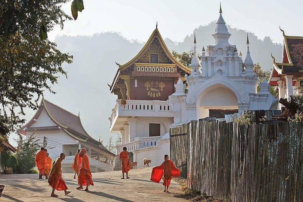 A imagem mostra um grupo de monges budistas vestidos com túnicas laranjas caminhando em uma rua tranquila. Ao fundo, há edifícios tradicionais com telhados ornamentados e uma estrutura branca que parece ser um templo. A paisagem é montanhosa, com árvores ao lado da estrada e uma luz suave do sol da manhã.