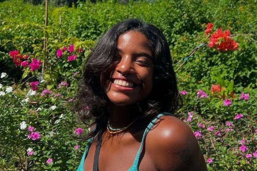 Uma mulher está sorrindo em um jardim cheio de flores coloridas. Ela tem cabelo solto e usa uma blusa de alças. O fundo é composto por várias plantas e flores, predominantemente rosas e outras cores vibrantes, criando um ambiente alegre e natural.