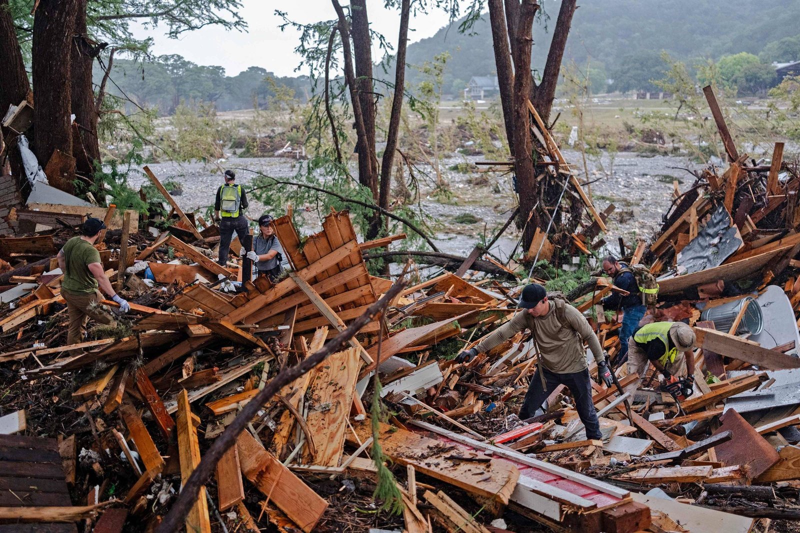 A imagem mostra um grupo de pessoas trabalhando em meio a destroços de madeira e outros materiais, possivelmente após um desastre natural. Há árvores ao fundo e uma área devastada, com sinais de destruição. As pessoas estão vestidas com roupas de trabalho e parecem estar realizando atividades de resgate ou limpeza.