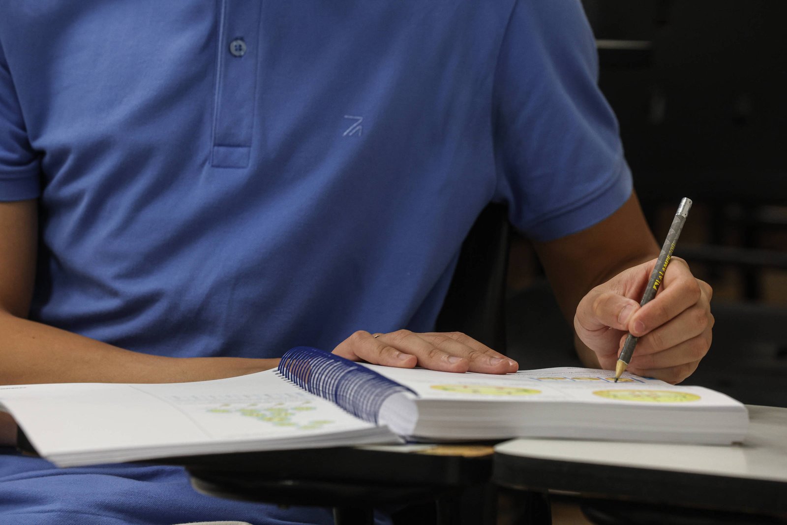 Um estudante está sentado em uma mesa de sala de aula, usando uma camisa azul. Ele está escrevendo em um caderno com uma caneta, enquanto uma folha de papel está aberta na mesa. O ambiente parece ser uma sala de aula, com cadeiras e mesas visíveis ao fundo.