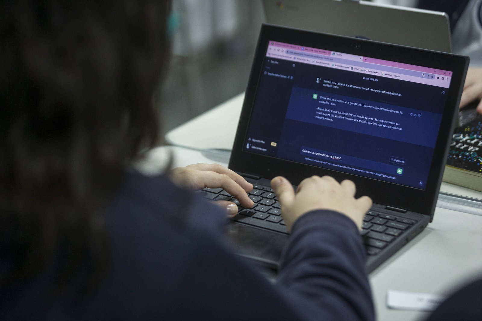 A imagem mostra uma pessoa de costas, com cabelo escuro, digitando em um laptop. A tela do laptop exibe uma interface de chat com fundo escuro e texto visível. Há uma mão de outra pessoa visível ao lado, sugerindo um ambiente colaborativo.