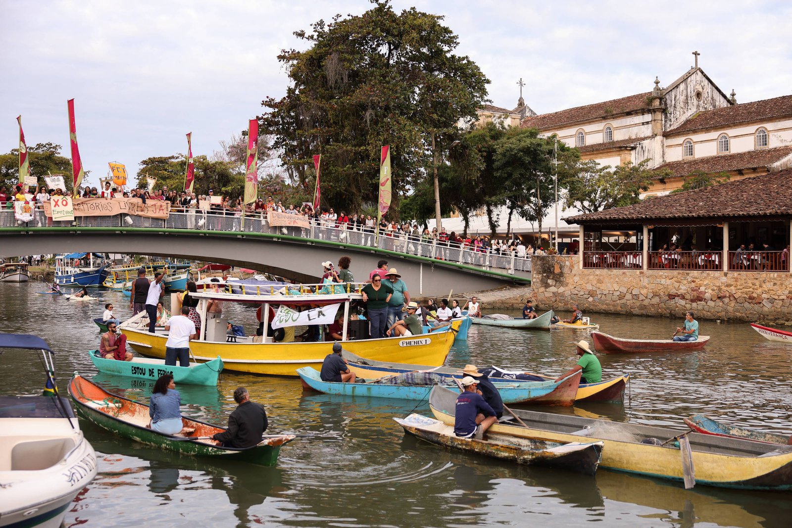 A imagem mostra várias embarcações coloridas navegando em um rio, com pessoas a bordo. Ao fundo, há uma ponte que conecta as duas margens do rio, e várias pessoas estão reunidas em uma área elevada, protestando com cartazes. O cenário é complementado por árvores e um edifício histórico visível ao fundo.