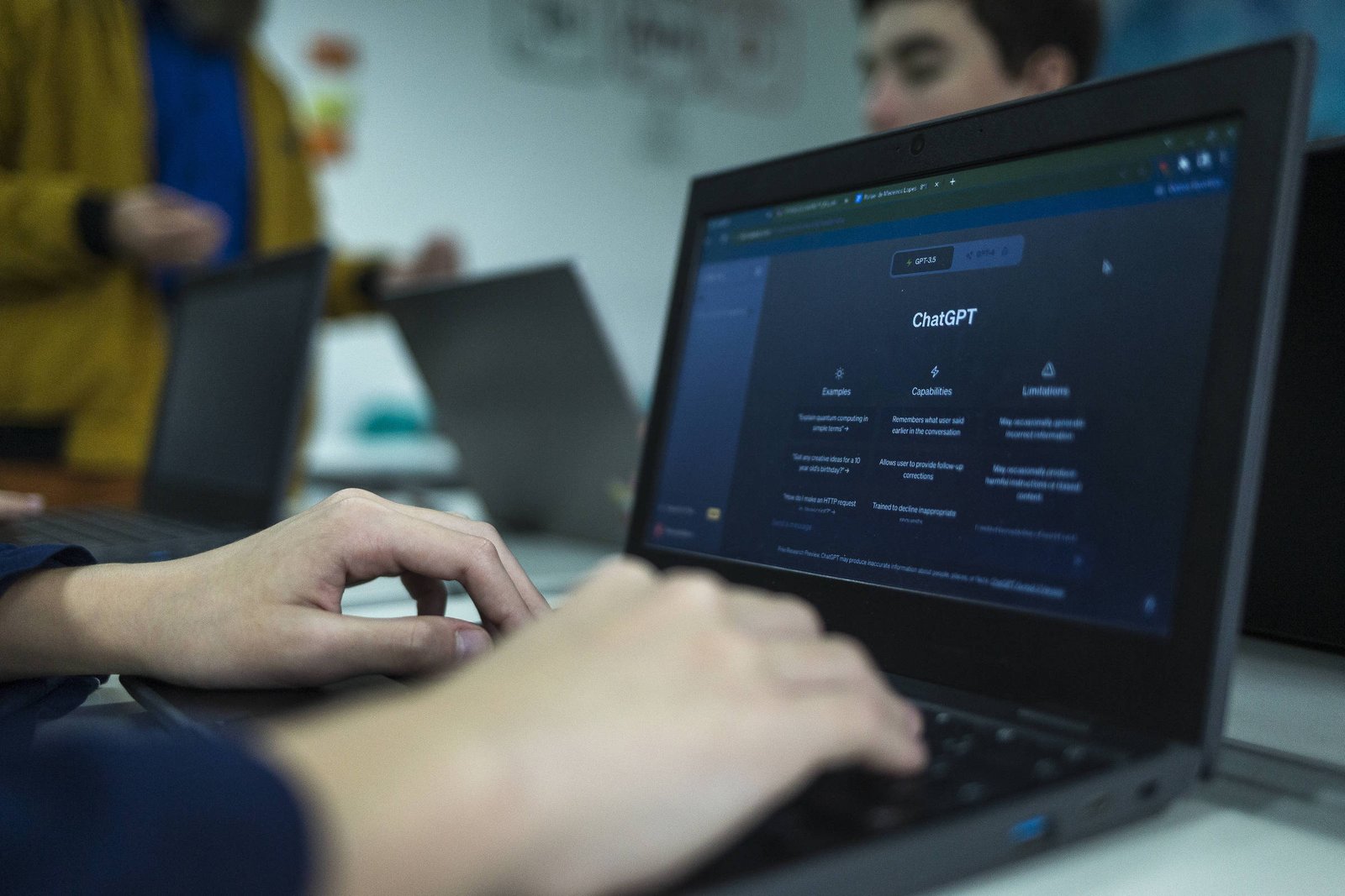 A imagem mostra uma pessoa digitando em um laptop que exibe a interface do ChatGPT. O fundo apresenta outras pessoas e laptops, com um ambiente de aprendizado ou trabalho. A tela do laptop é escura e contém seções sobre