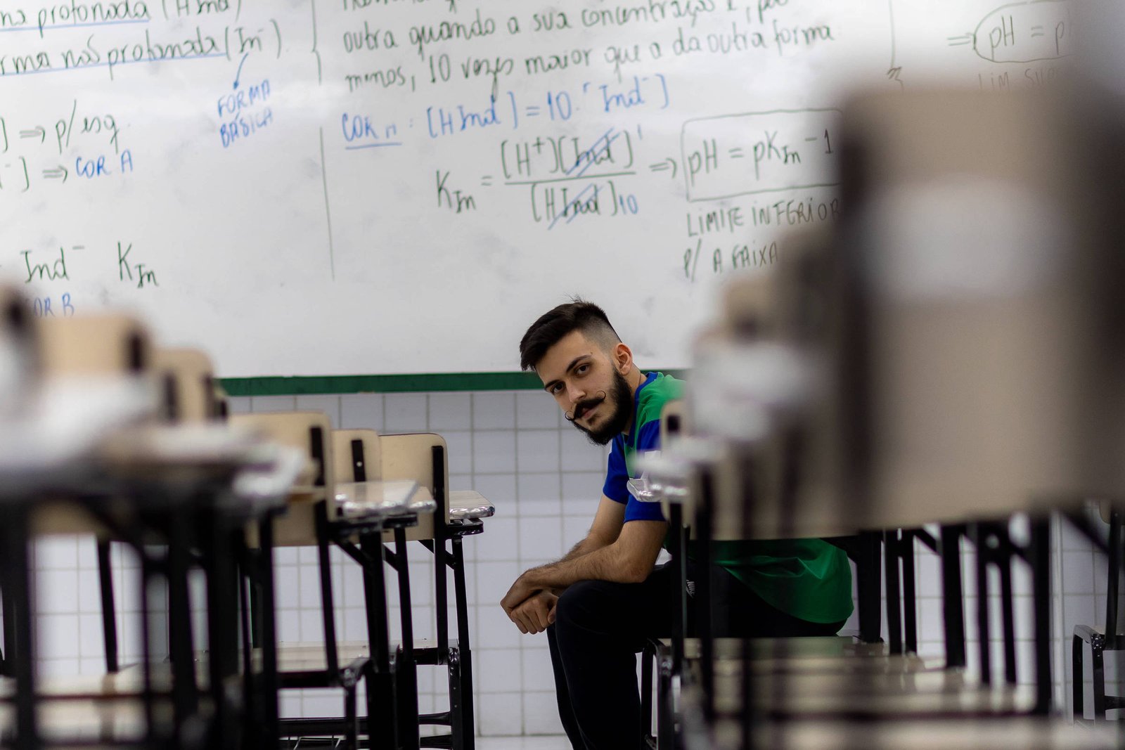 Um jovem sentado no chão de uma sala de aula, entre as mesas e cadeiras. Ele tem cabelo curto e barba, vestindo uma camiseta azul e um colete verde. Ao fundo, uma lousa branca com anotações e fórmulas escritas em caneta preta e azul. A sala parece vazia, com as mesas organizadas em fileiras.