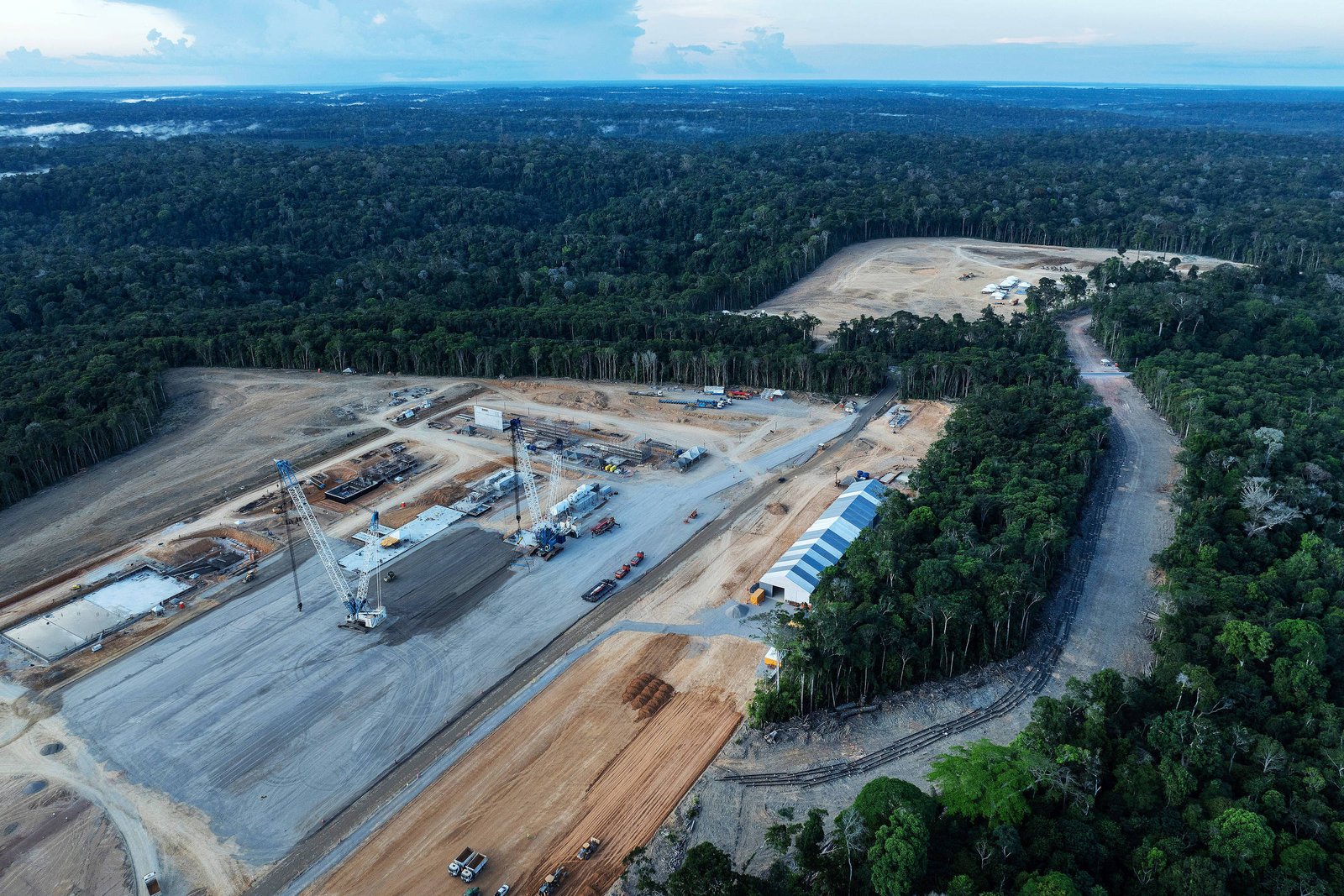A imagem mostra uma área de construção em uma região florestal. No primeiro plano, há uma grande área desmatada com maquinário e estruturas de construção visíveis. Ao fundo, a floresta densa se estende até o horizonte, com árvores altas e uma paisagem verde. O céu está parcialmente nublado, e há uma estrada que se afasta da área de construção.