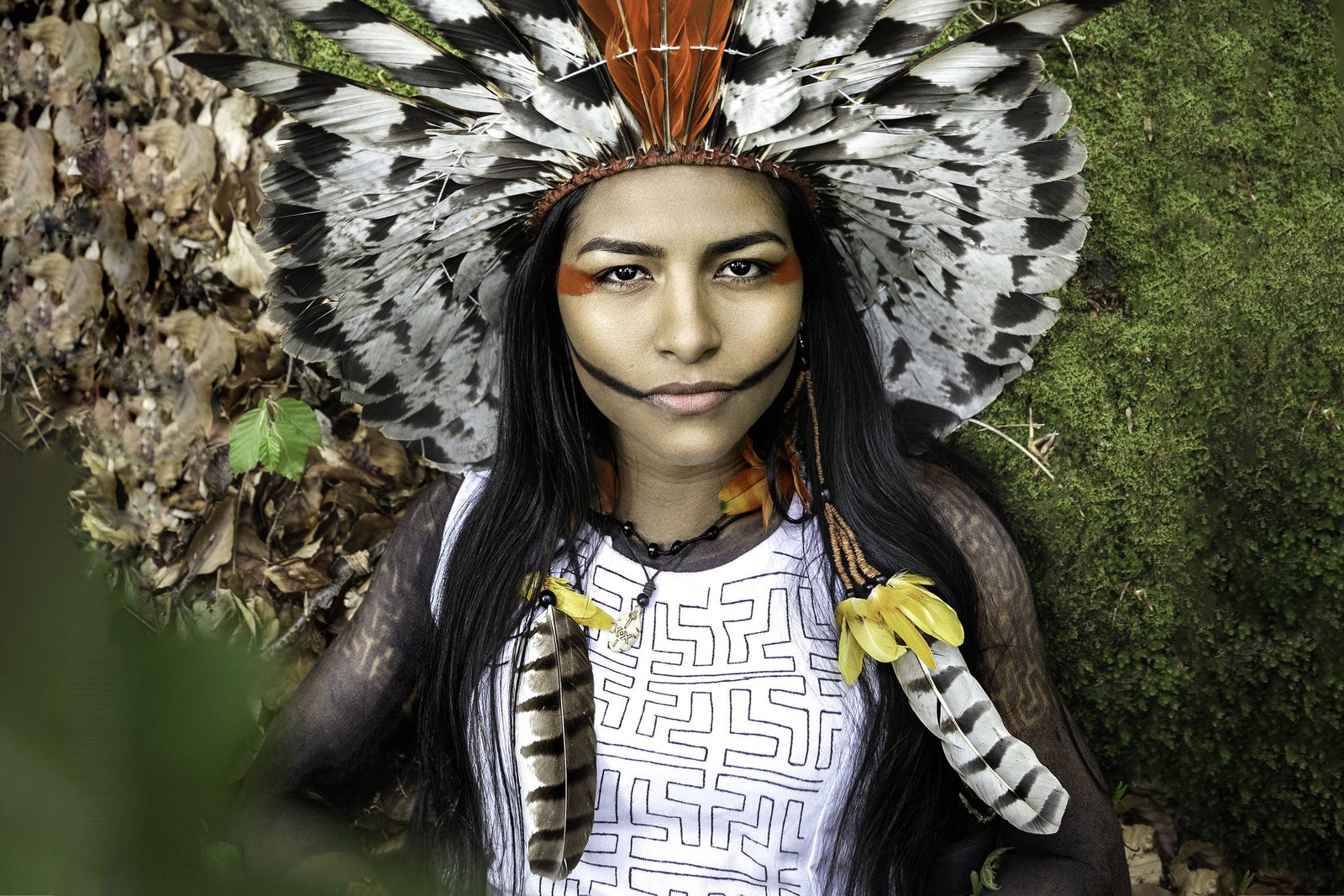 A imagem mostra uma jovem indígena de rosto sereno, usando um elaborado cocar de penas. Ela está em frente a um fundo natural com folhas secas e musgo. Seu rosto é pintado com cores vibrantes, incluindo laranja e preto, e ela usa colares com penas e outros adornos. A iluminação é suave, destacando os detalhes do cocar e da maquiagem.