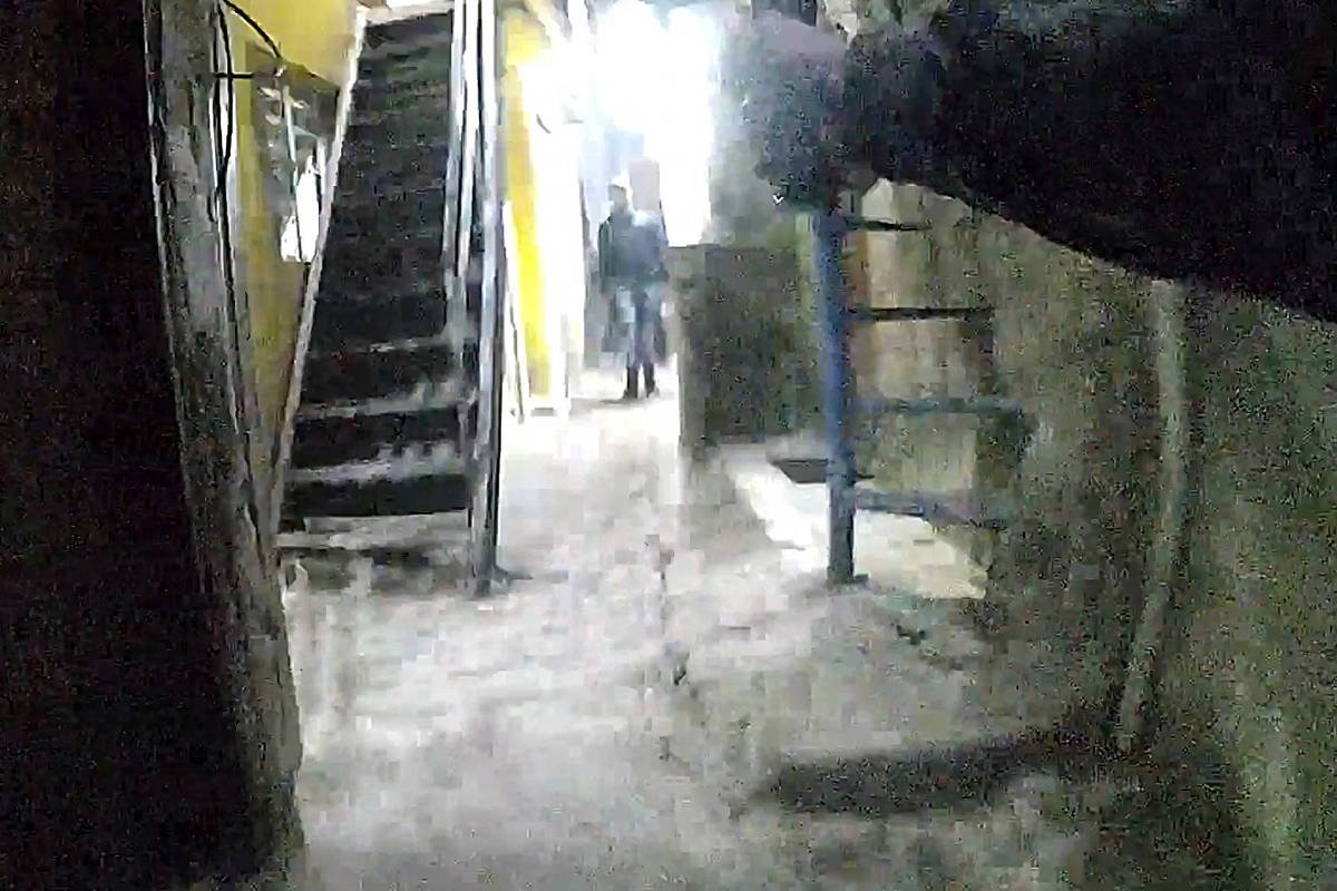 A imagem mostra um corredor estreito com escadas à esquerda. O chão é de cimento. À direita, há uma parede e uma escada azul. No fundo, uma pessoa está de pé, parcialmente visível, em um ambiente iluminado por uma luz branca. As paredes são de cor clara.